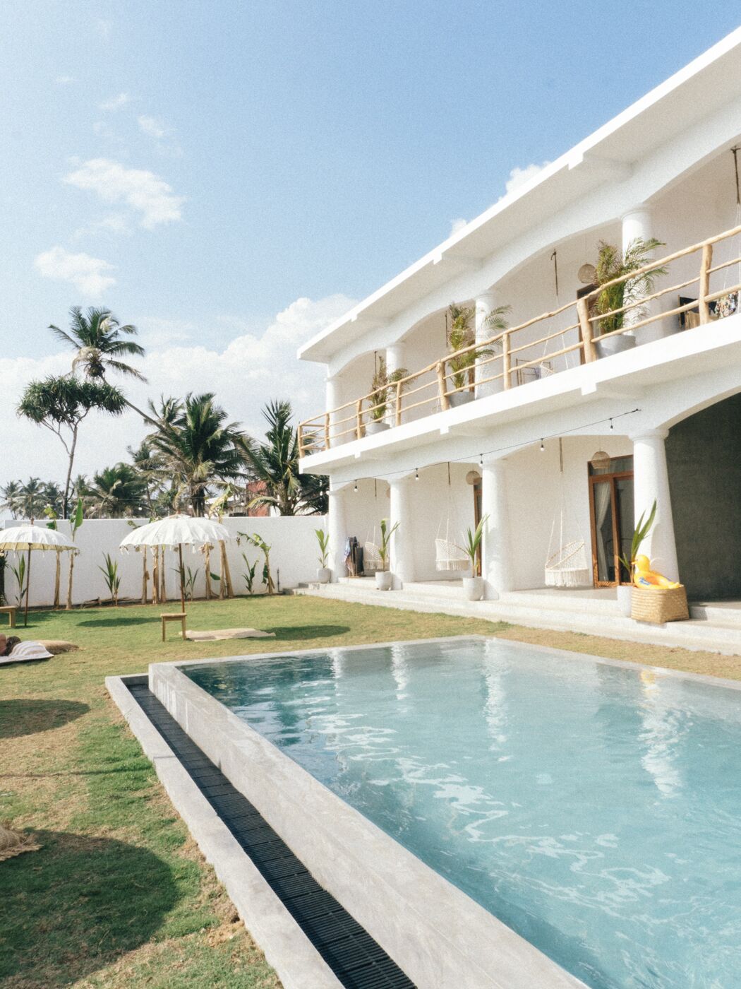 Un área de piscina con una piscina azul cristalina, un edificio blanco con balcón y palmeras al fondo.