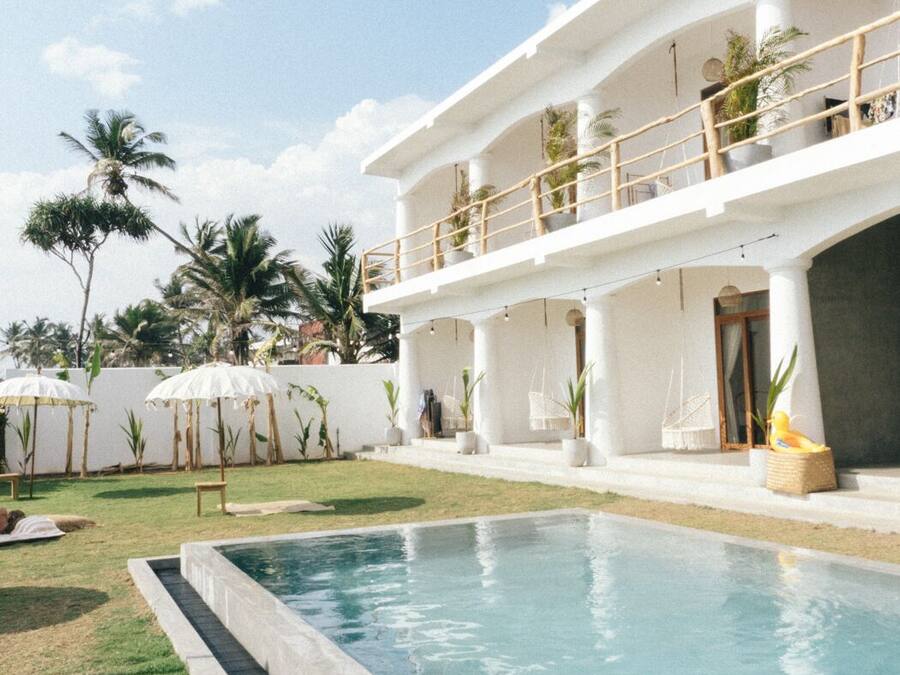 A pool area with a clear blue pool, a white building with a balcony, and palm trees in the background.