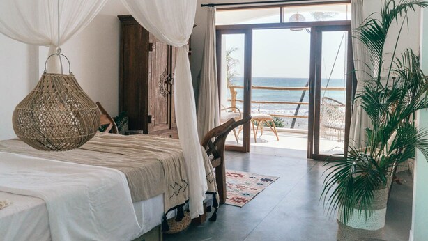 Une chambre avec un lit, une lampe suspendue, une vue sur la plage et une grande plante en pot.