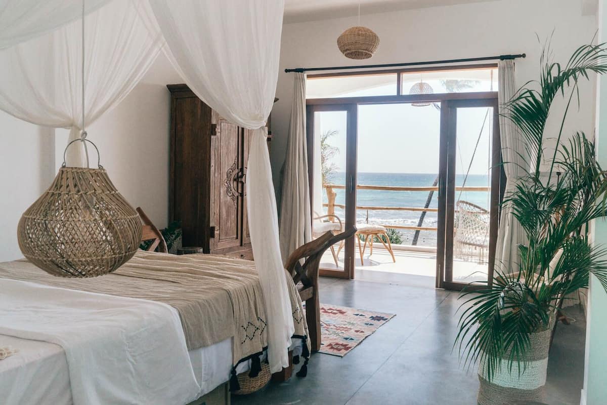 Een slaapkamer met een bed, een hanglamp, uitzicht op het strand en een grote kamerplant.