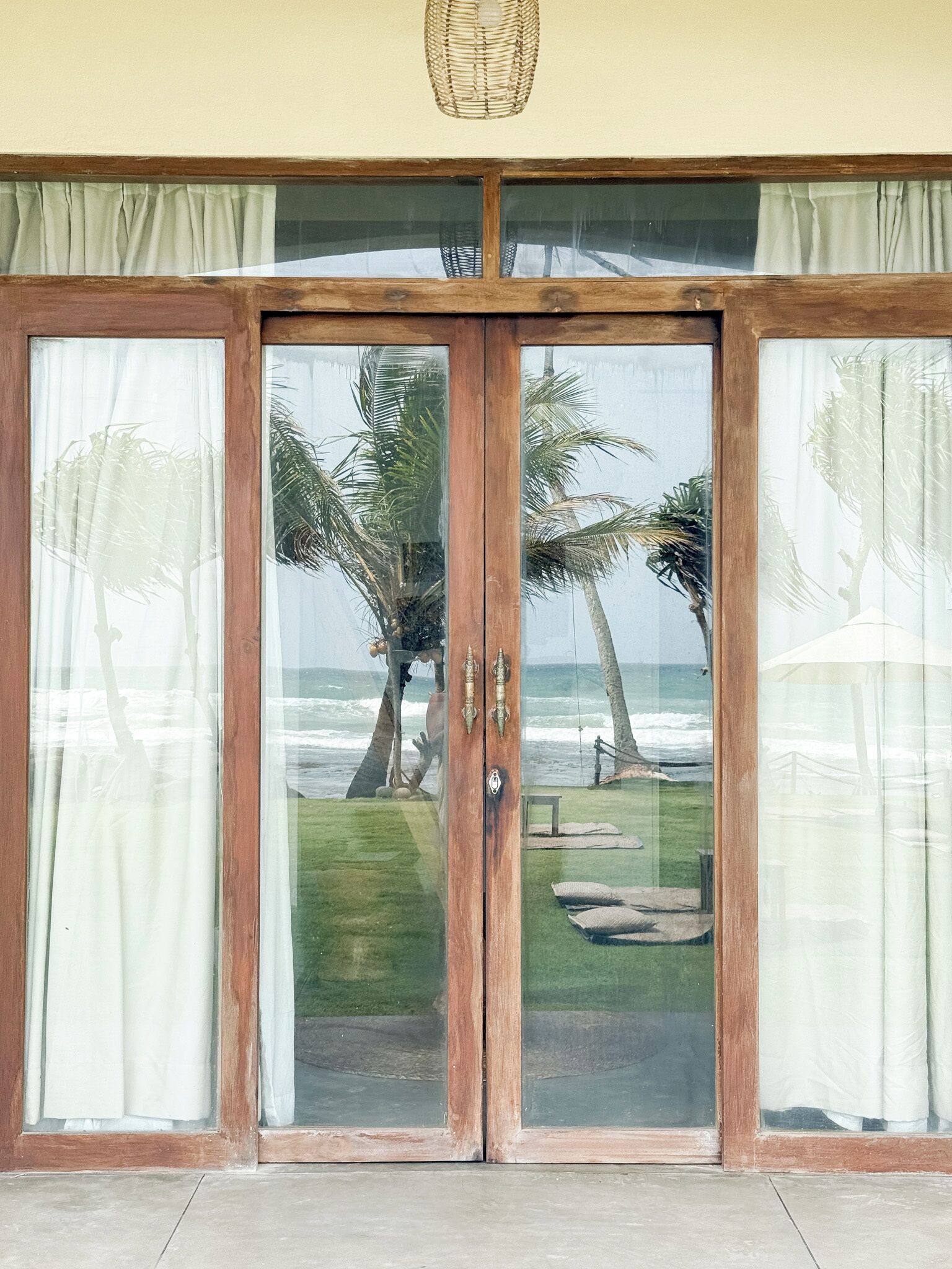 A wooden door with glass panels, leading to a view of the ocean and palm trees.
