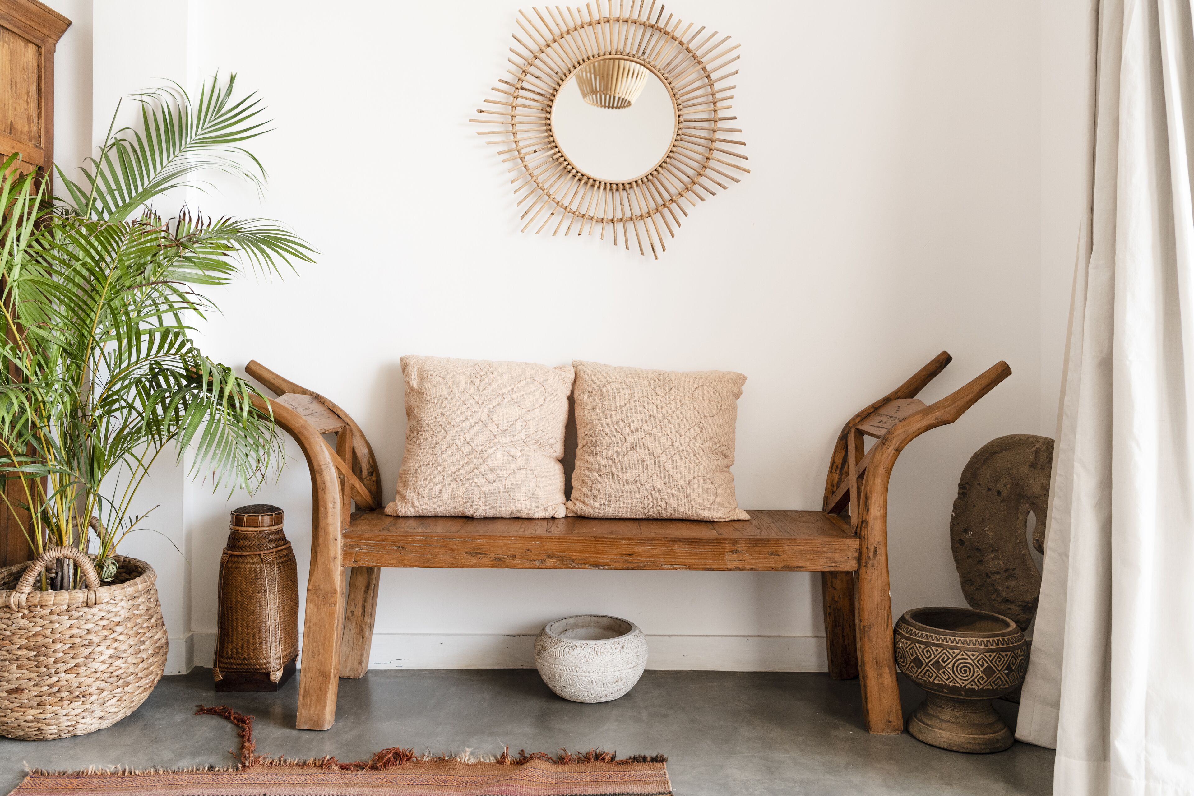 Een houten bank met twee kussens, een rieten mand, een potplant en een decoratief wandobject.