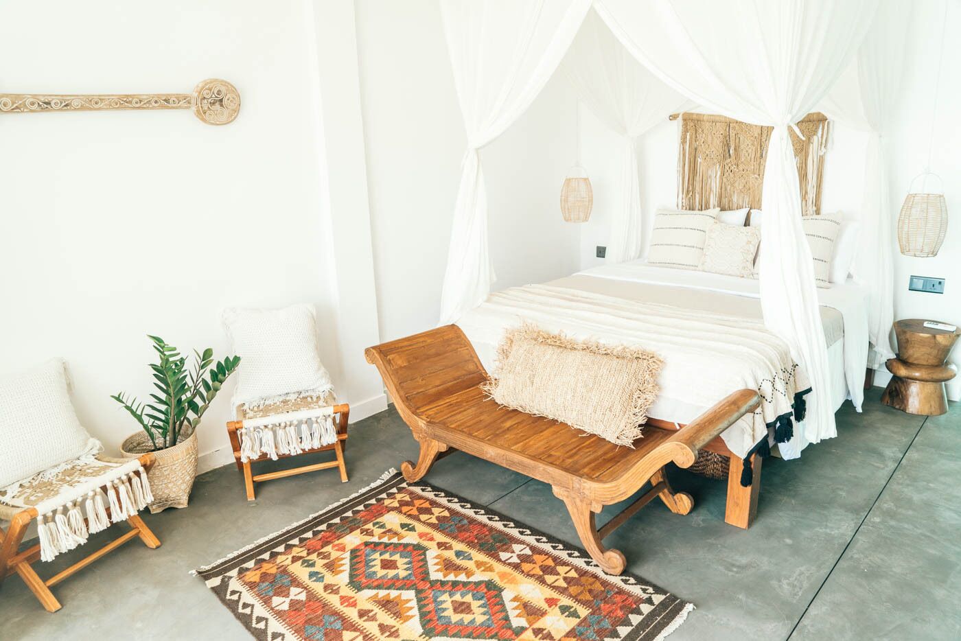 A bedroom with a canopy bed, a wooden bench, a patterned rug, and a plant.