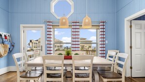 A dining area with a large table, white chairs, and a view of houses through a window with striped curtains. - New! Rare Mezzanine Deck, Hot Tub & Beach Views! (Crystal Beach)