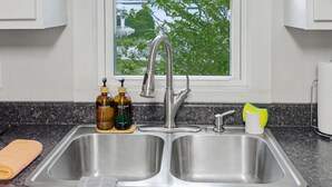 Una cocina con un lavamanos de acero inoxidable doble, dos botellas de jabón, una esponja amarilla y una ventana con follaje a la vista.