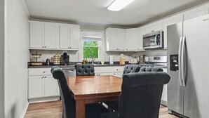 Una cocina moderna con armarios blancos, una mesa de comedor de madera, sillas tapizadas en negro, un refrigerador de acero inoxidable y una ventana con vistas a zonas verdes.