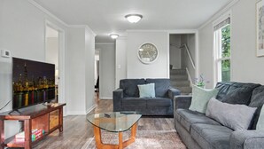 Una sala de estar con televisión de pantalla plana, dos sofás, una mesa de centro de vidrio y un reloj en la pared.