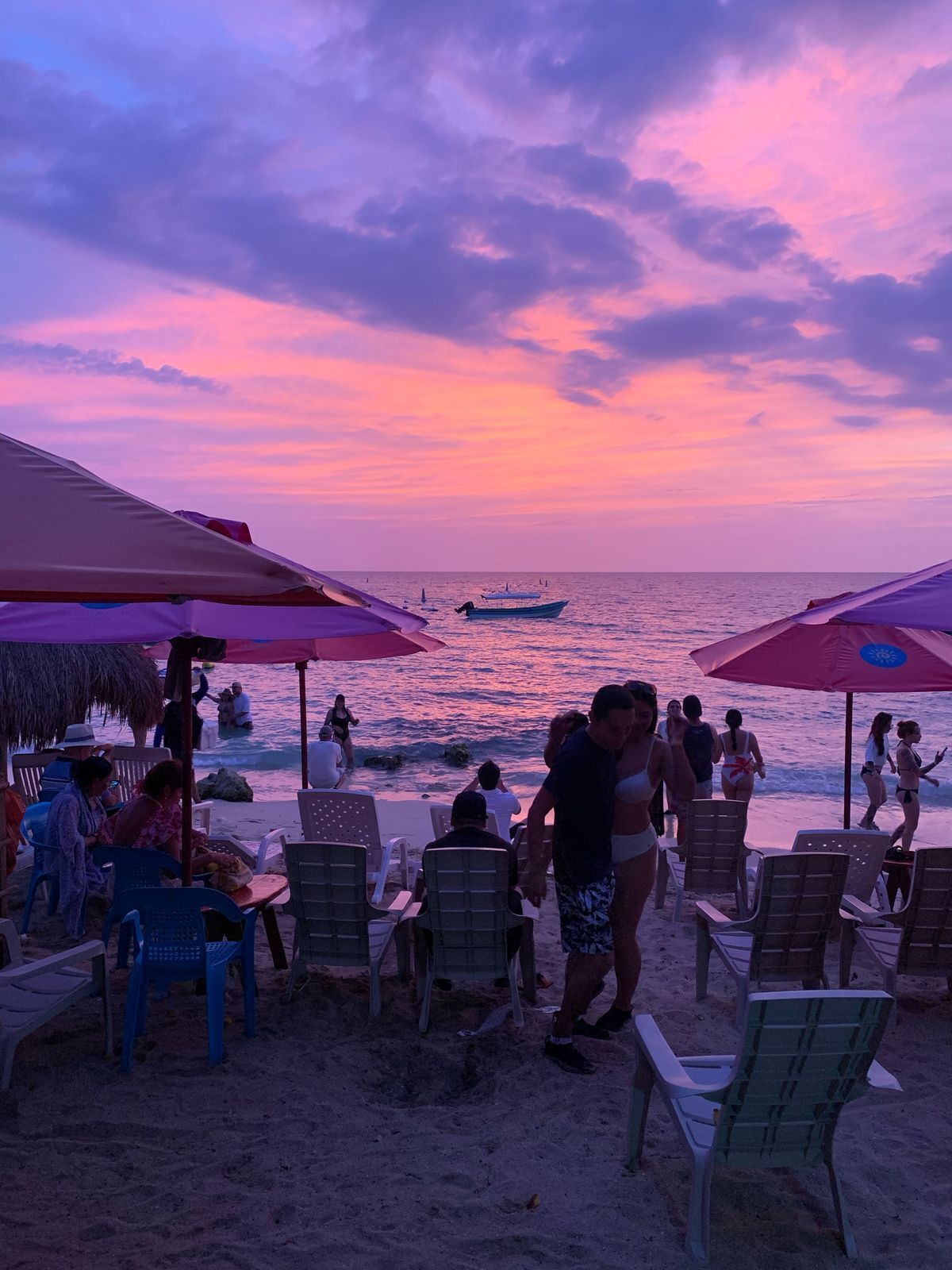 A beachside scene with people relaxing under umbrellas, a boat in the water, and a sunset in the background.