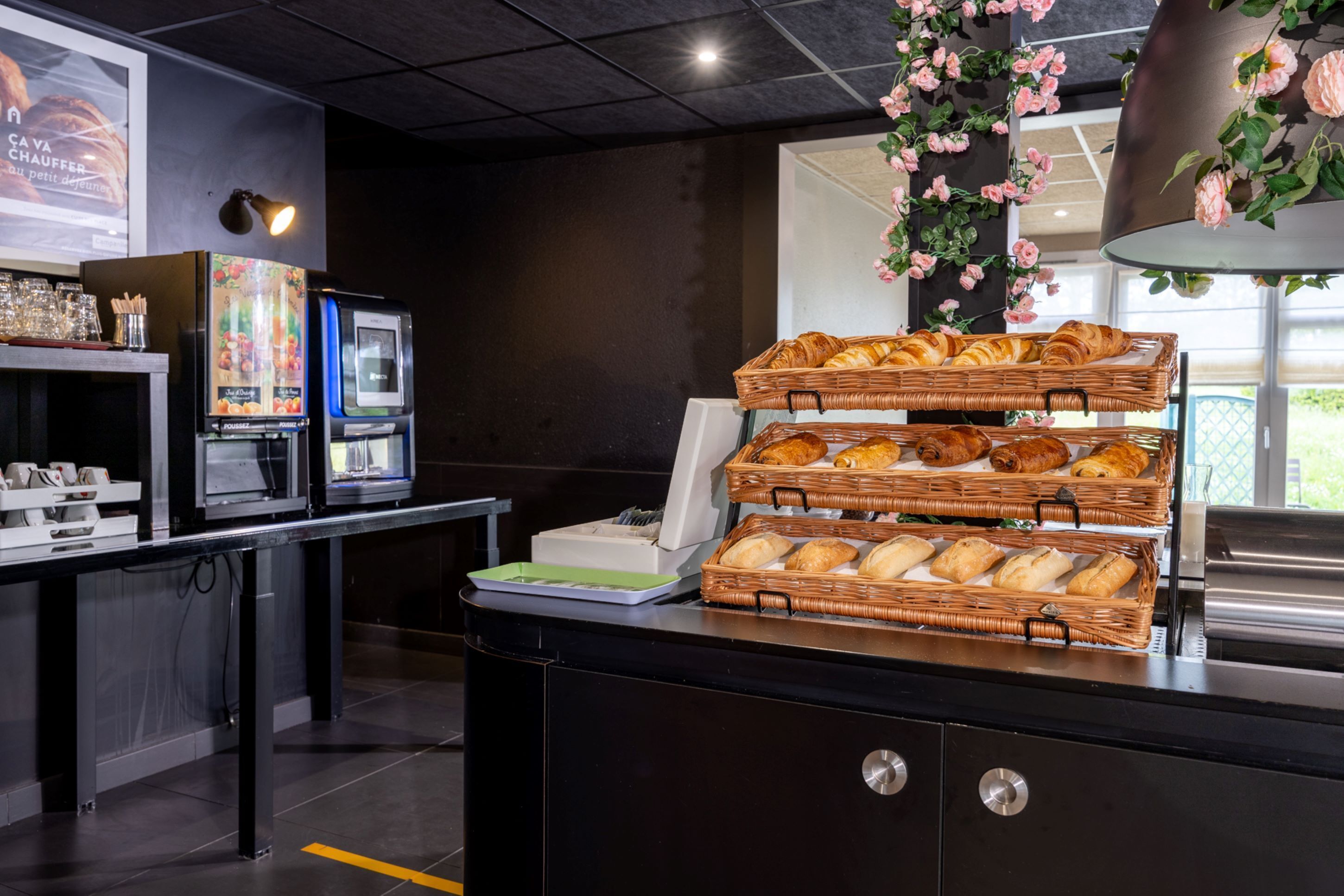 A breakfast buffet with wicker baskets of croissants and bread, a coffee machine, and a beverage dispenser.