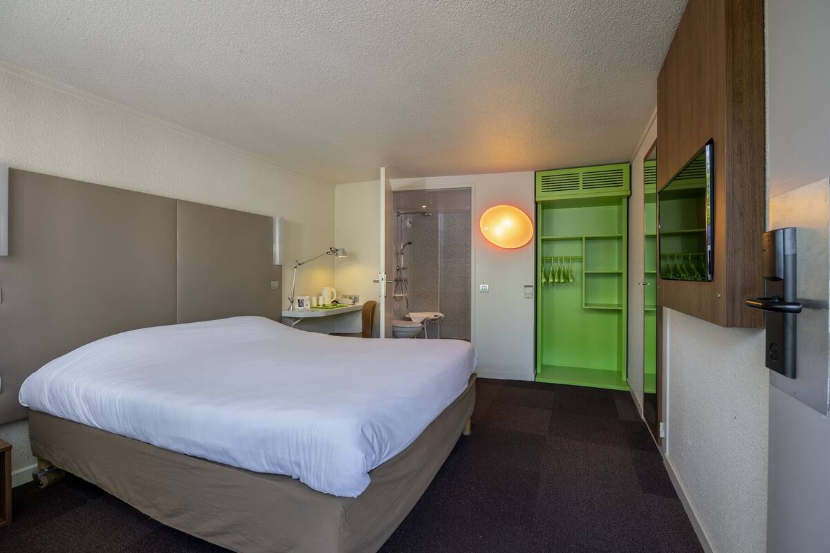 A hotel room with a bed, a desk with a lamp, a shower, and a green wardrobe.