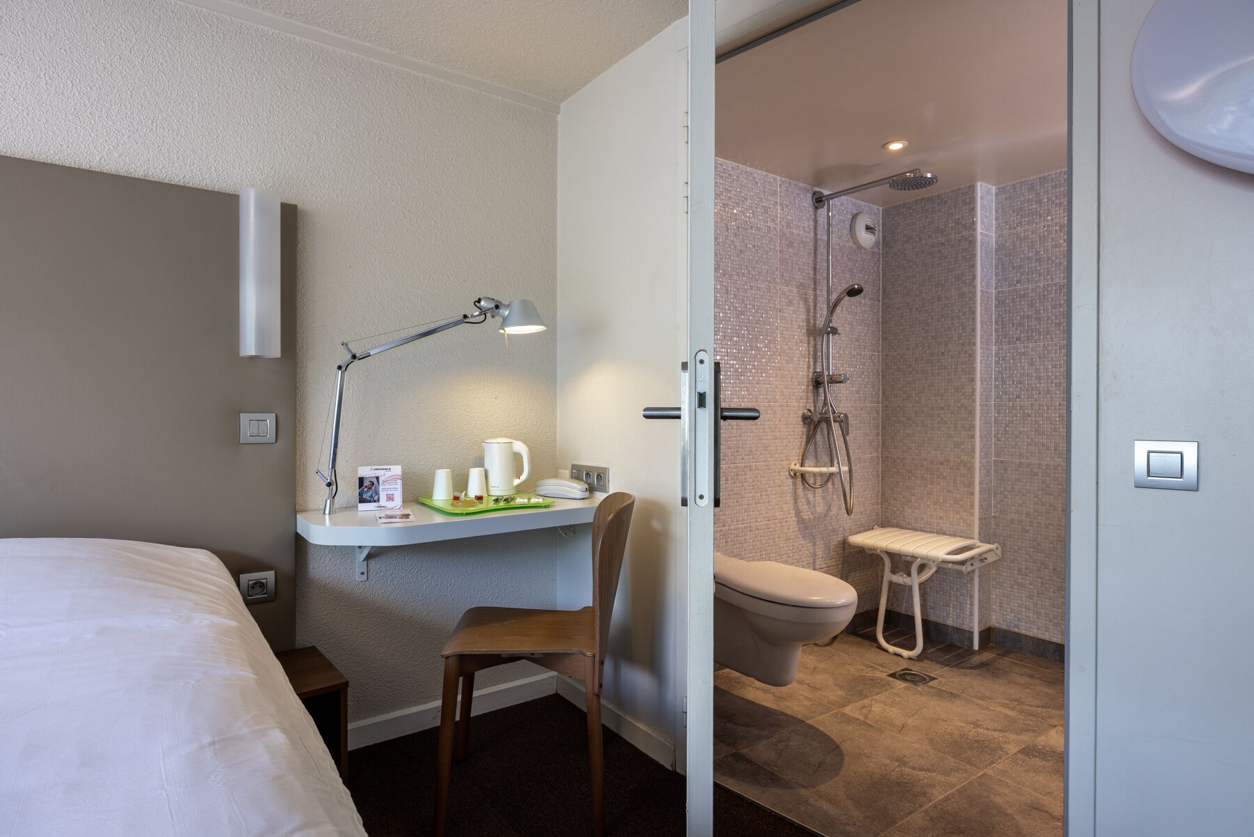 A hotel room with a bed, a desk with a lamp, a chair, a small table with a mug, a shower, and a toilet.
