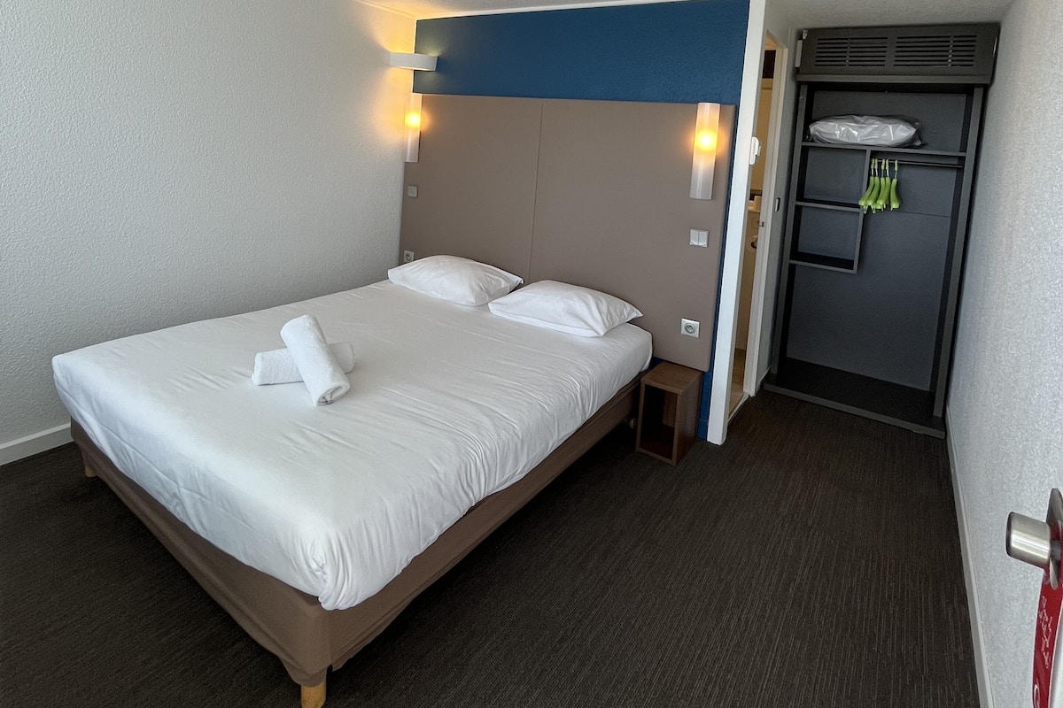 A hotel room with a single bed, a headboard with wall-mounted lights, a small bedside table, and a built-in wardrobe with a mirror.