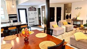 A modern kitchen with white cabinets, a dining area with a wooden table and chairs, and a living room with a sofa and armchairs. - Chef's Kitchen + Minutes to Boyne Mountain + Walk Downtown (Boyne City)