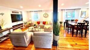 A modern living room with a sofa set, a dining table with chairs, a TV mounted on the wall, and a large window. - Chef's Kitchen + Minutes to Boyne Mountain + Walk Downtown (Boyne City)