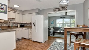 A modern kitchen with white cabinets, a refrigerator, and a dining area with a wooden table and chairs. - 4BR Home w/ Private Dock and Fenced Backyard and Firepit. Minutes from the Beach (St. Augustine)
