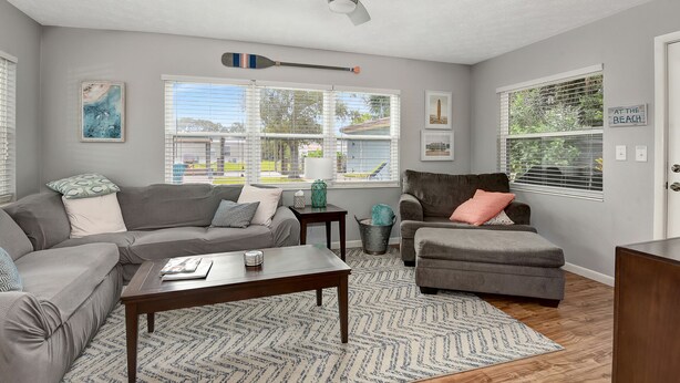 A living room with a gray sofa, a wooden coffee table, and a gray armchair. There are two windows with blinds, a ceiling fan, and a wall-mounted paddleboard. - 4BR Home w/ Private Dock and Fenced Backyard and Firepit. Minutes from the Beach (St. Augustine)