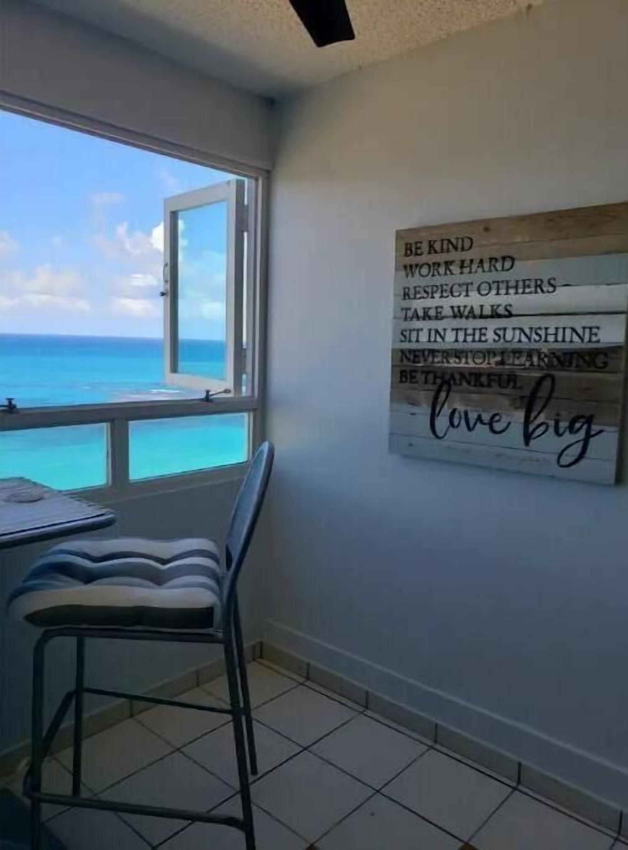 A room with a view of the ocean, a chair, a table, and a motivational quote on the wall.
