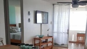 A spacious living area with a wooden dining table, cushioned chairs, a TV on the wall, and a large window with curtains. - Luquillo beachfront apt., beautiful ocean view! (Luquillo)