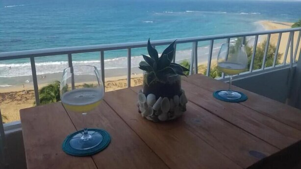 A wooden table with two wine glasses and a pineapple on a balcony overlooking a beach and ocean. - Luquillo beachfront apt., beautiful ocean view! (Luquillo)
