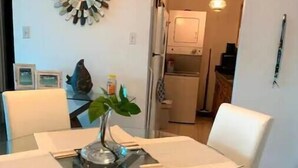 Un comedor con una mesa dispuesta para cuatro personas, rodeada de sillas blancas. Hay una planta en maceta sobre la mesa y un adorno decorativo en la pared superior.