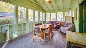 Une véranda abritée avec une table et des chaises en bois, un ventilateur de plafond et une vue sur les arbres à travers de grandes fenêtres.