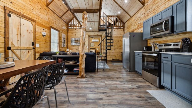 Interior de una cabaña de madera con comedor, cocina y una escalera que sube a una buhardilla.