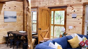A wooden cabin interior with a dining table, chairs, a sofa, and a door leading outside. - 2 Cabins- Hot Tubs | Dogs OK | Hiking | Climbing (Campton)