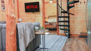 A cozy room with a wooden interior, a bed, a sofa, a small table with a plant, and a TV mounted on the wall displaying "NETFLIX". - 2 Cabins- Hot Tubs | Dogs OK | Hiking | Climbing (Campton)