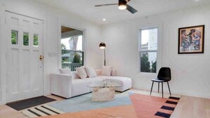 A modern living room with a white sofa, a glass coffee table, and a black chair. There is a large window with blinds, a ceiling fan, and a framed picture on the wall. - Charming Plymouth retreat • hot tub • fire pit

 (Plymouth)