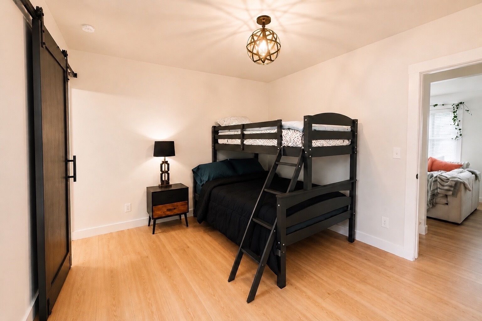 A bedroom with a bunk bed, a bedside table with a lamp, and a door leading to another room.