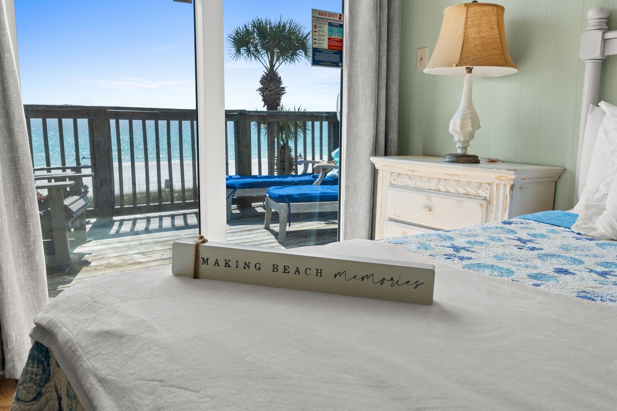 A hotel room with a view of the beach, a bed with a blue and white patterned bedspread, a bedside table with a lamp, and a sign on the window reading 'Making Beach Memories'.