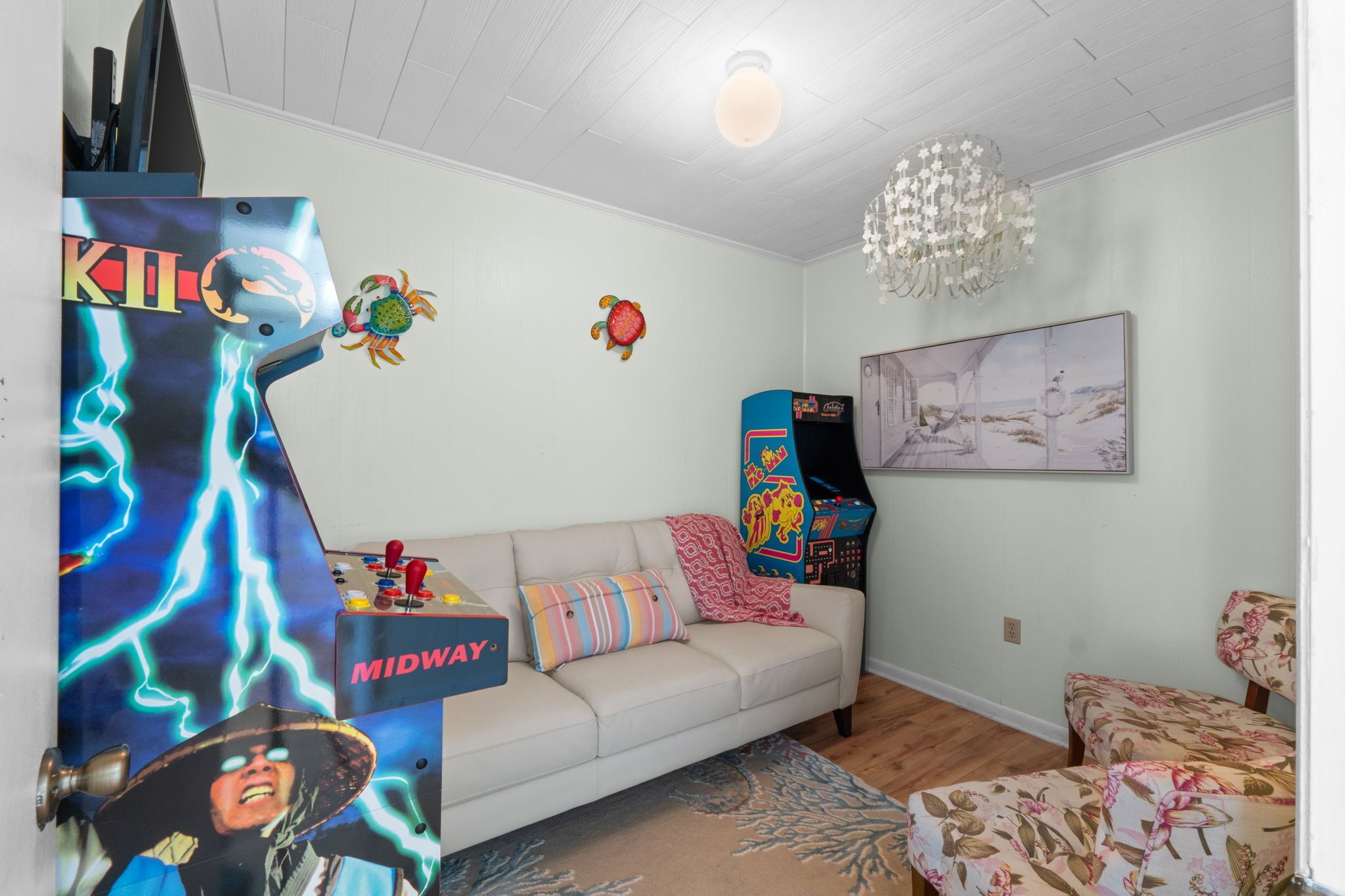 Una habitación con techo blanco, un candelabro, un sofá, una máquina de videojuegos y un cuadro en la pared.