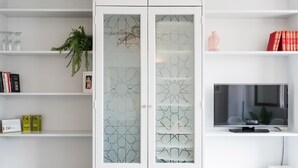 A modern living room with white cabinetry, a glass-fronted cabinet with geometric patterns, a flat-screen TV, and a potted plant. - Alhambra View Suite Apartamentos Bravo (Granada)
