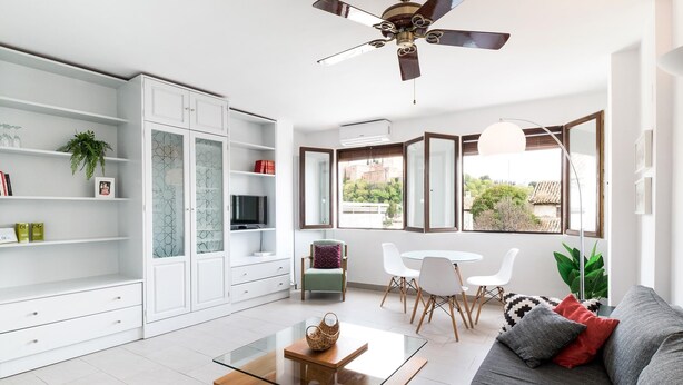 A modern living room with a glass coffee table, a sofa, a dining area with a white table and chairs, and a large window with a view of buildings and greenery. - Alhambra View Suite Apartamentos Bravo (Granada)