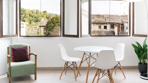 A dining area with a round table and four white chairs, a green armchair with a patterned cushion, and a potted plant. - Alhambra View Suite Apartamentos Bravo (Granada)