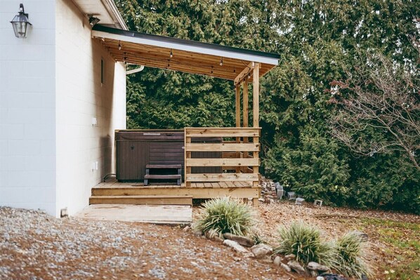 Una terraza de madera con un jacuzzi y una mesita pequeña, que da a un edificio blanco con una pérgola.