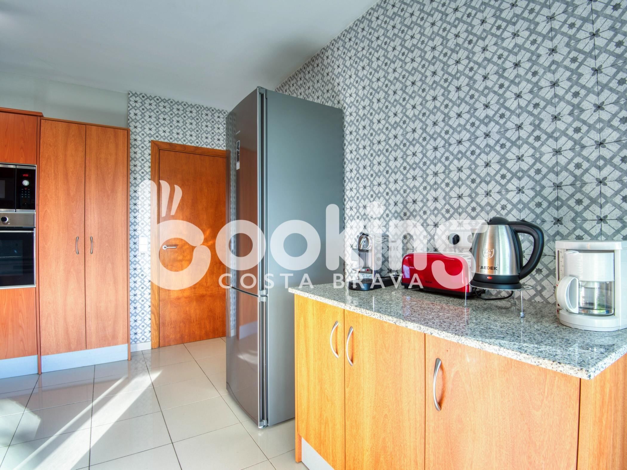 A kitchen with wooden cabinets, a stainless steel refrigerator, a coffee maker, and a kettle on the countertop.