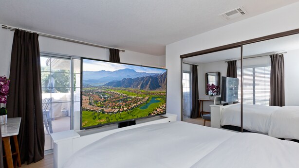 A modern hotel room with a large bed, a flat-screen TV displaying a scenic view, a desk with a chair, and a large window with curtains. - Los Balcones de Palm Springs Mid-Century Modern Designer Condo, City ID #068484 (Palm Springs)