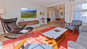 A modern living room with a large flat-screen TV, a dining area with a table and chairs, and a view of the outdoors through a large window. - Los Balcones de Palm Springs Mid-Century Modern Designer Condo, City ID #068484 (Palm Springs)