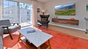 A modern living room with a large window, a sofa, a coffee table, a chair, and a TV wall-mounted above a wooden cabinet. - Los Balcones de Palm Springs Mid-Century Modern Designer Condo, City ID #068484 (Palm Springs)