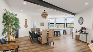 A modern kitchen with a dining area, featuring a large window, a hanging light fixture, and a bamboo plant.