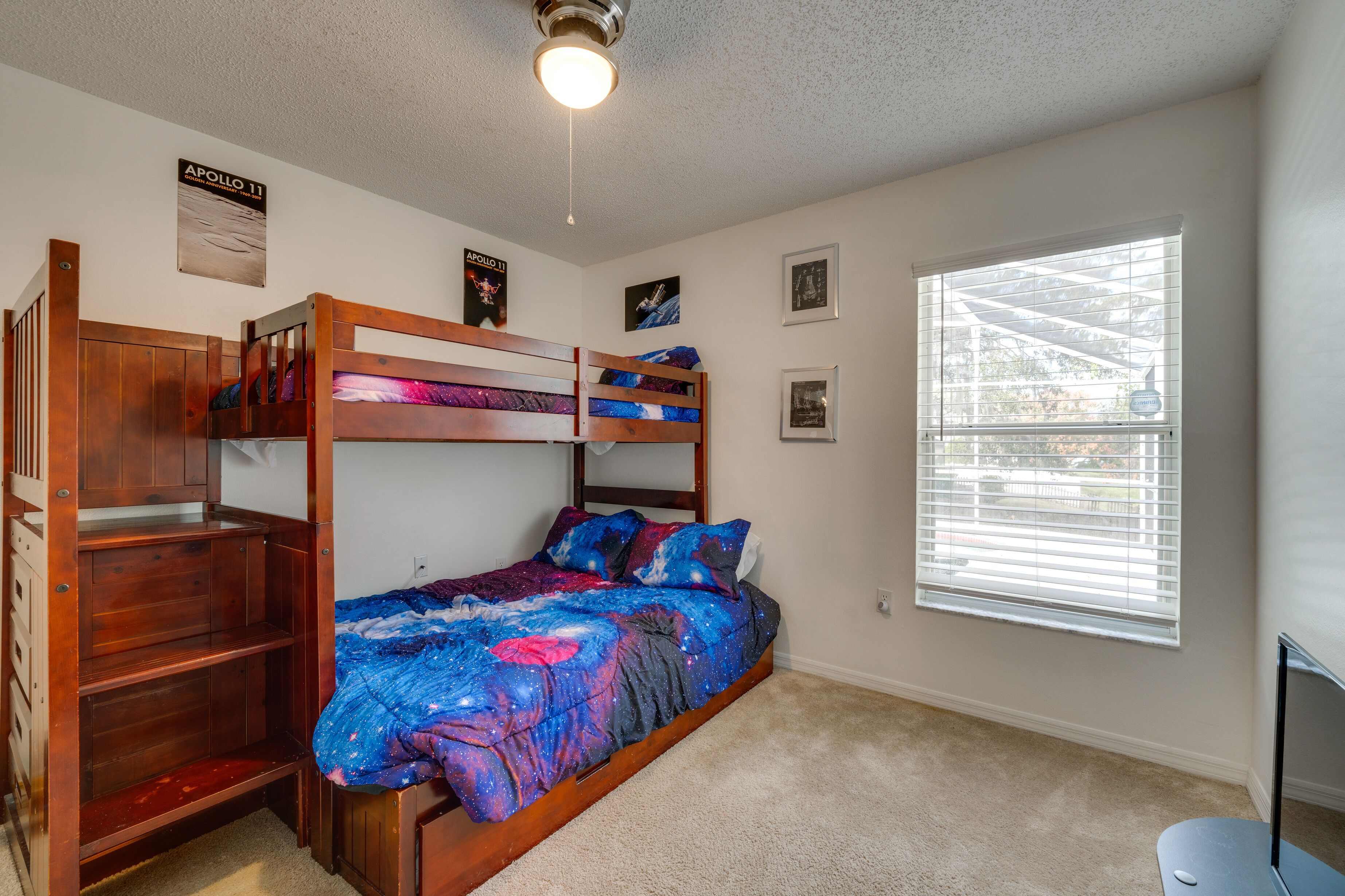 A bunk bed room with a galaxy-themed bedspread, a wooden desk, and a window with blinds.
