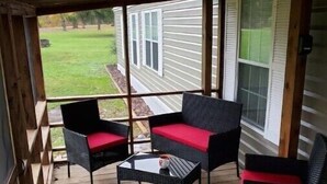 Un espacio cubierto para sentarse al aire libre con muebles de mimbre y cojines rojos, con vistas a un jardín césped y una casa.