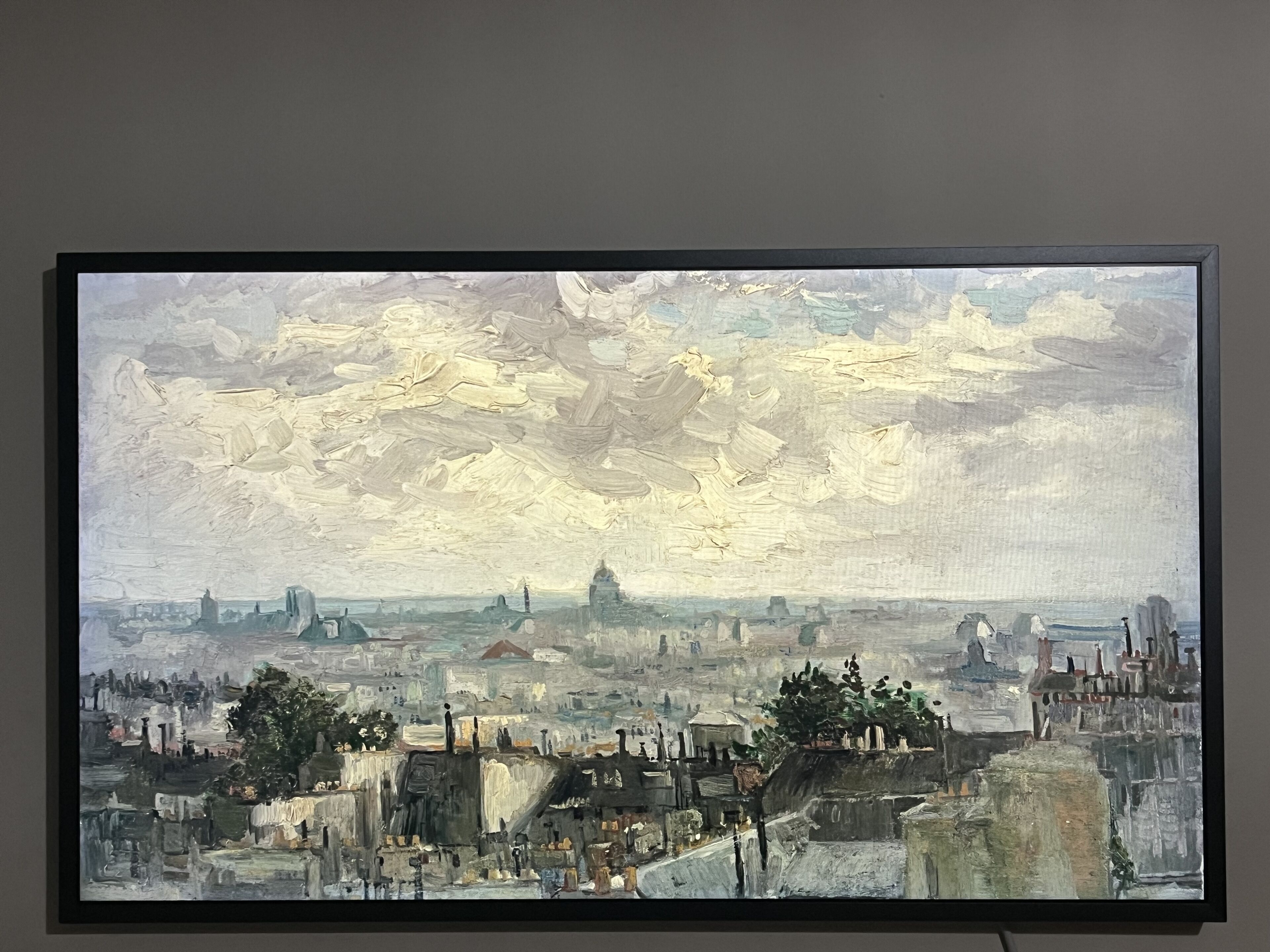 Ein gerahmtes Gemälde einer Stadtansicht mit Gebäuden, Bäumen und einem bewölkten Himmel.