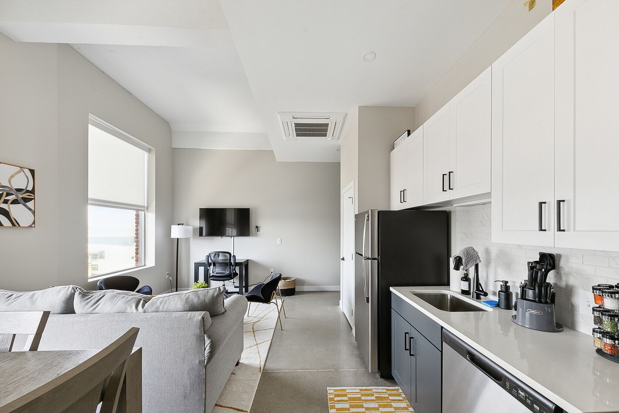 A modern kitchen with white cabinets, a black refrigerator, and a stainless steel sink. There is a dining area with a wooden table and chairs, and a gray sofa. A television is mounted on the wall, and a window provides natural light.
