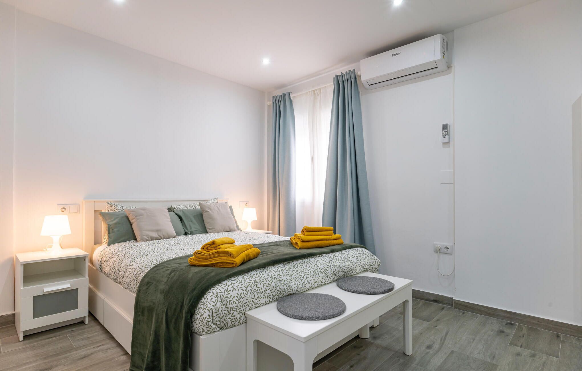 A modern bedroom with a bed, bedside tables, a bench, and an air conditioning unit.