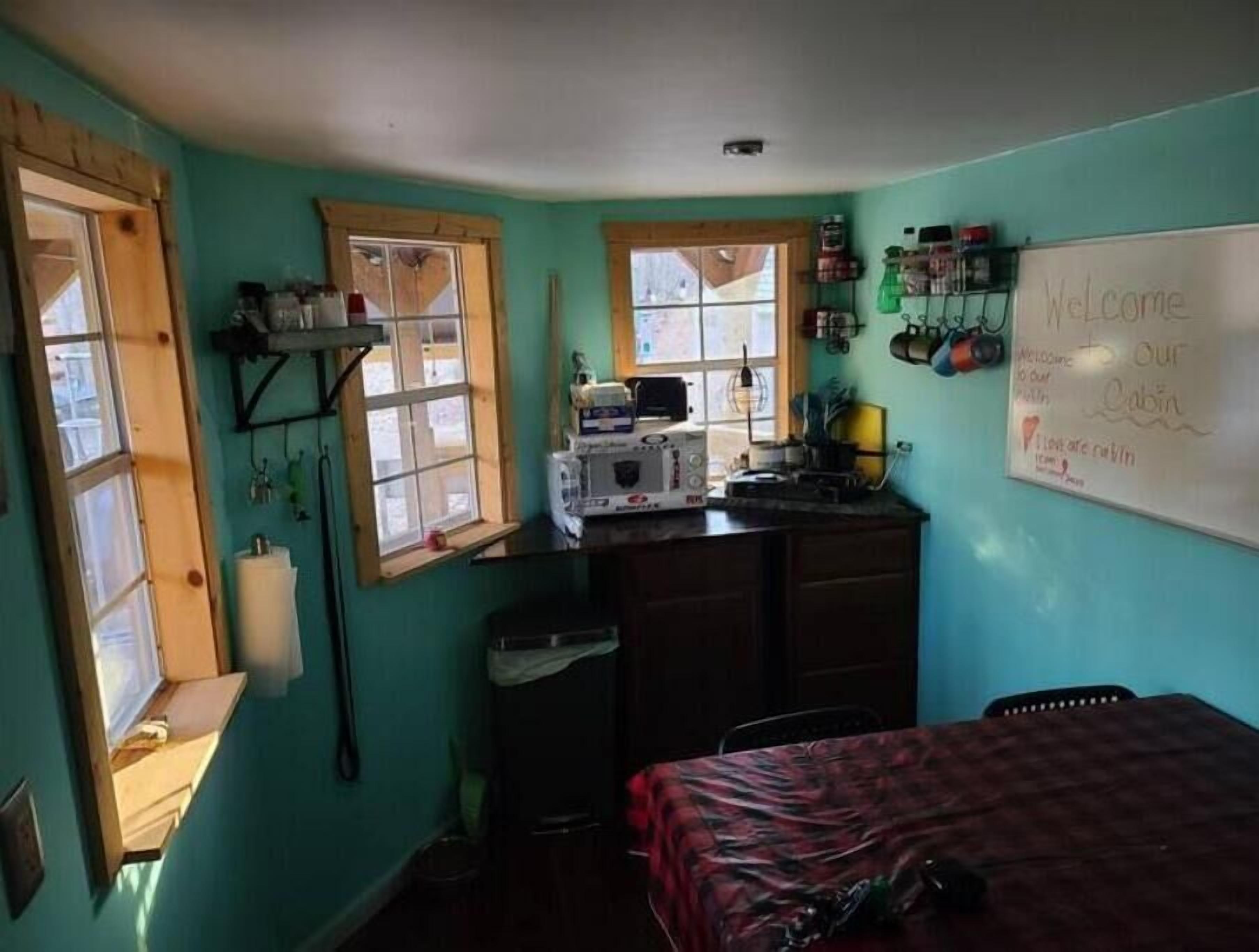 A small, cozy room with teal walls, a wooden desk with a sewing machine, a whiteboard with a welcome message, and a bed with a checkered bedspread.