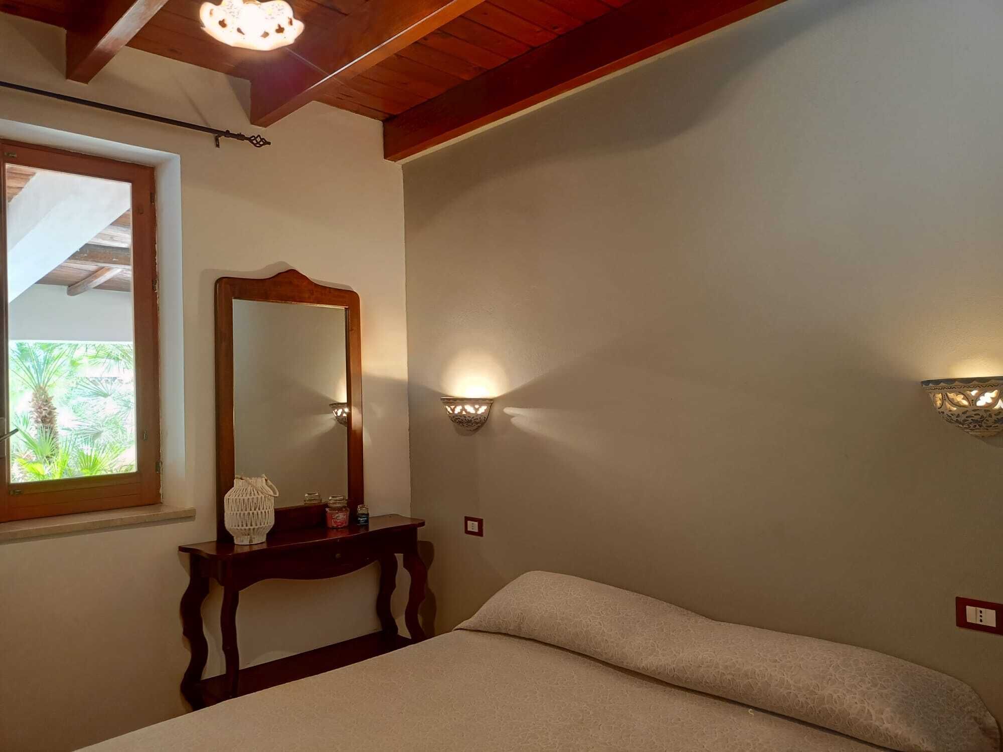 Quarto com teto de madeira, espelho grande, luminárias de cabeceira e janela com vista para área verde.
