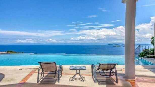 Zona piscina con due sedie e un tavolo, con vista su un mare blu cristallino e isole in lontananza.