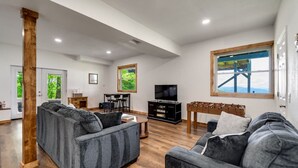 Un salon spacieux avec un grand canapé gris, une table basse en bois et un téléviseur posé sur un meuble noir. On y trouve également une table en bois devant laquelle se trouve un baby-foot, le tout placé devant une fenêtre donnant sur un paysage verdoyant.